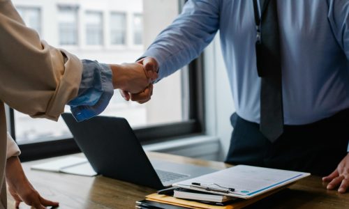 Crop unrecognizable coworkers in formal wear standing at table with laptop and documents while greeting each other before meeting